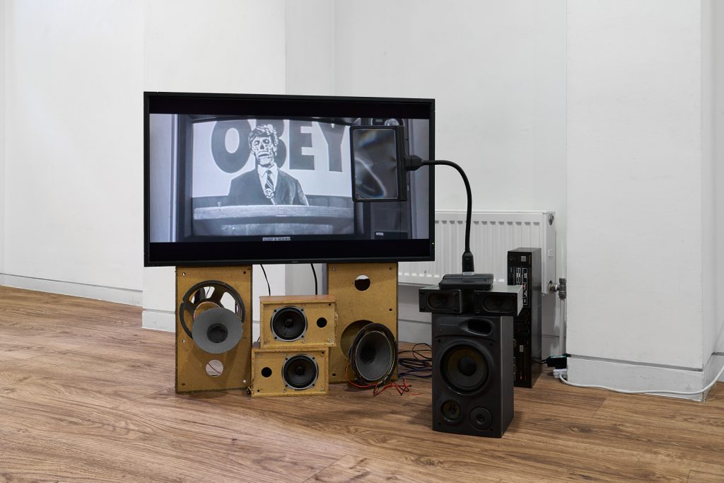 A flatscreen monitor sits on top of some broken or deconstructed analogue speakers. The image on the screen is a still from John Carpenter's film 'They Live'.