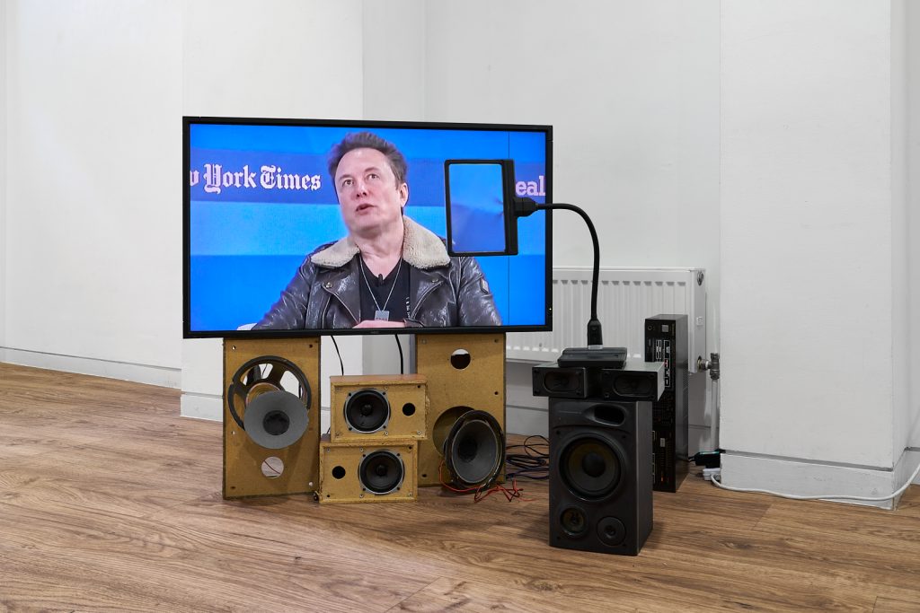 A flatscreen monitor sits on top of some broken or deconstructed analogue speakers. The image on the screen is a still image of Elon Musk speaking at an unknown event.