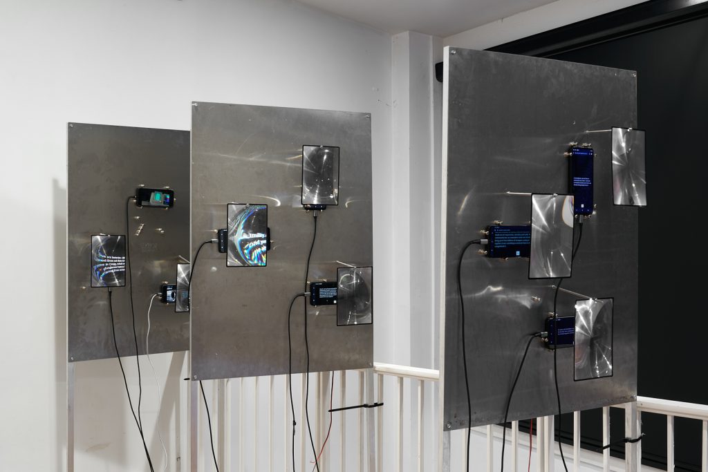Three metal panels are fixed to freestanding frames, each panel has three smartphones fixed to it. Text appears on the screens of the phones. Magnifiers hover over the screens, distorting the appearance of the text.