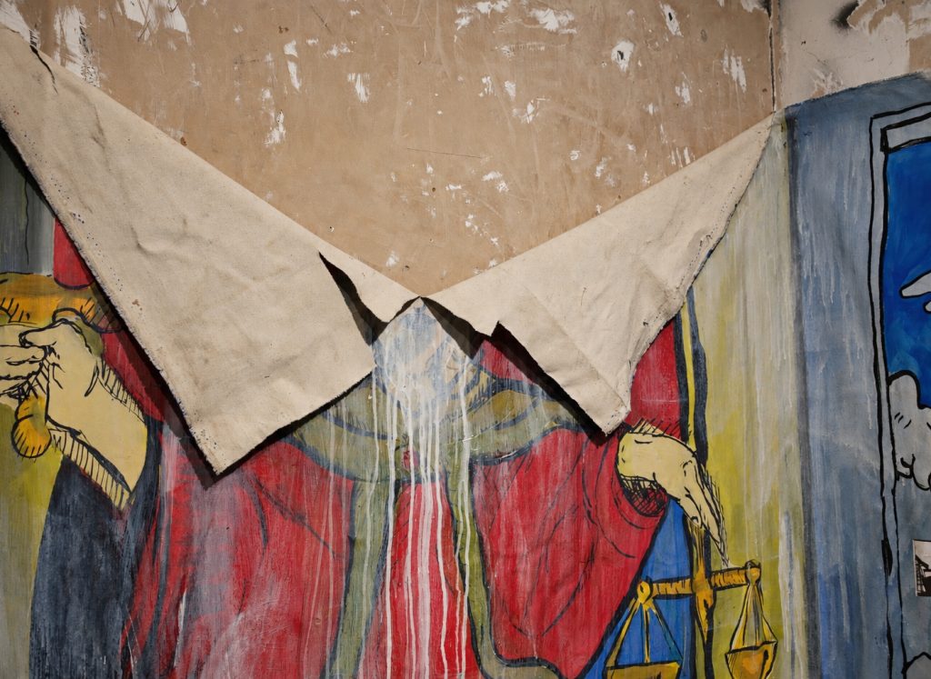 A torn painting on canvas of the Justice tarot, splashed with white paint, installed on a bare plaster wall
