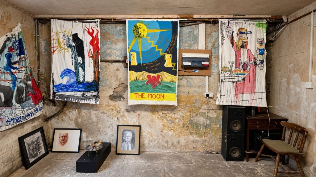 Painted banners showing re-versioned tarot cards hang along a basement wall. Along the foot of the wall are arranged framed images of famous people and a ceramic work on a low black plinth