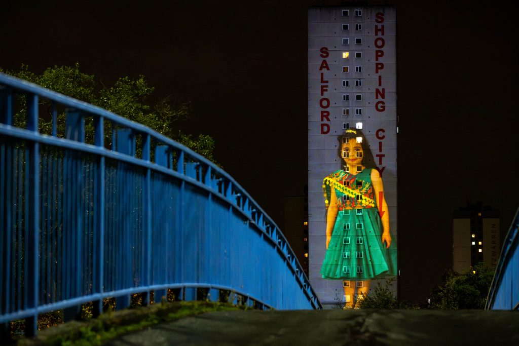 An image of a young girl in a bright green and yellow dress is projected onto the side of a tall building