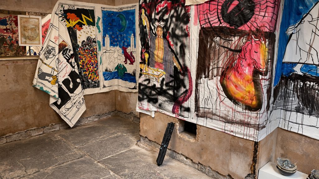 Painted banners showing re-versioned tarot cards hang along a basement wall. Along the foot of the wall are arranged dark ceramic works