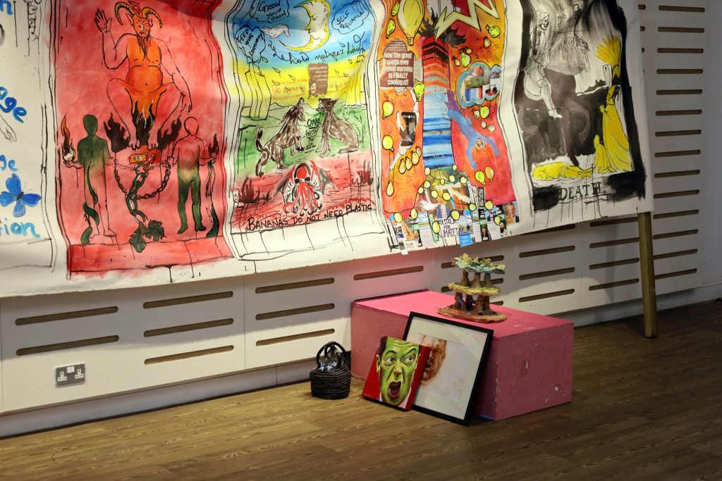 Painted banners installed in a gallery space; in the foreground, a white plinth with ceramic works and paintings arranged around and on top