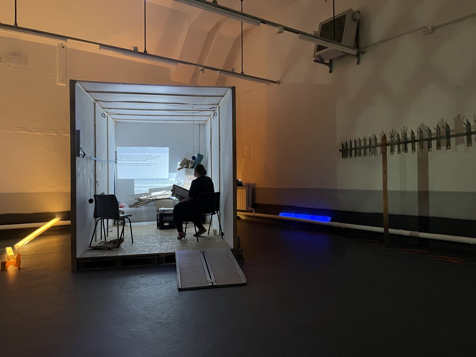 A container in a dimly lit space. Inside a person sits on a chair. There are images, like screen shots, projected on the interior of the container.