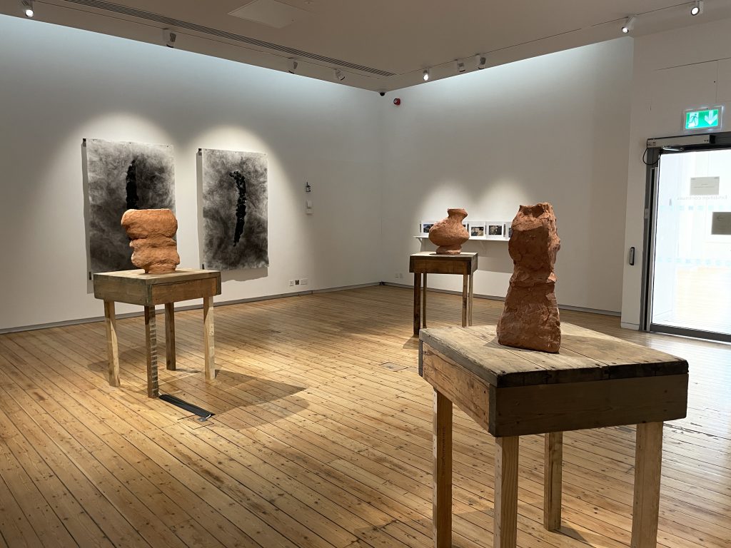 Three roughly hewn tables, each with a terracotta object on. Behind on the wall two large charcoal drawings.