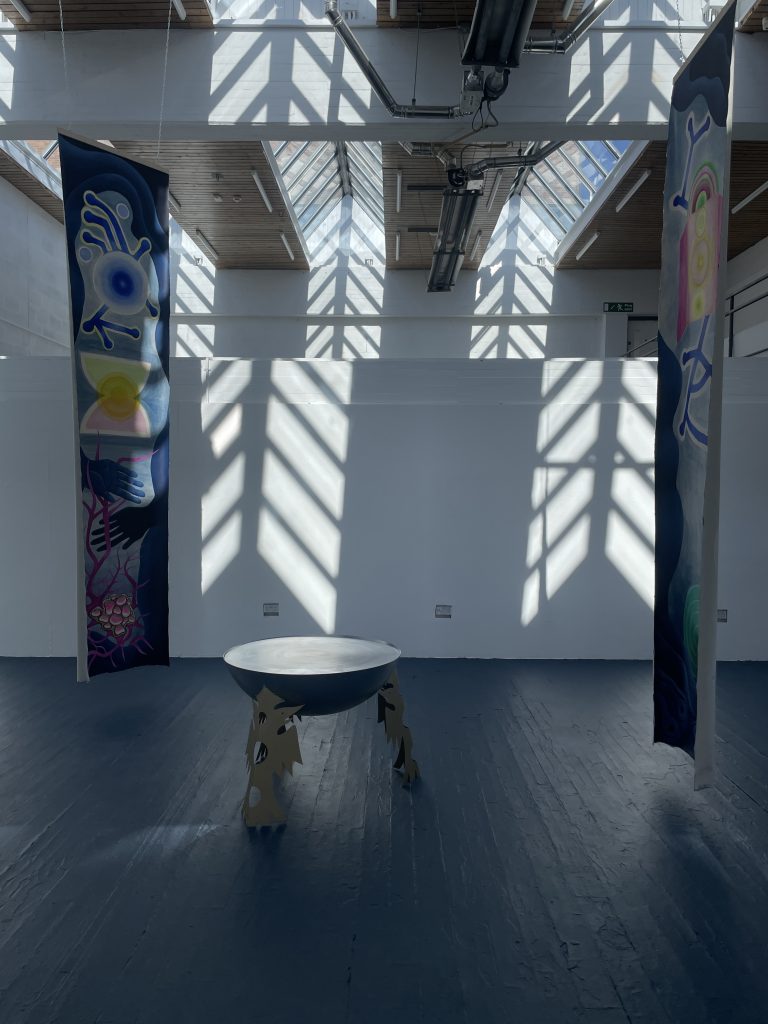 A room lit by daylight, filtered through skylights creating an effect on the surfaces. Two banners hang from the ceiling, painted with colourful designs. On the floor between them a font or large bowl on legs.