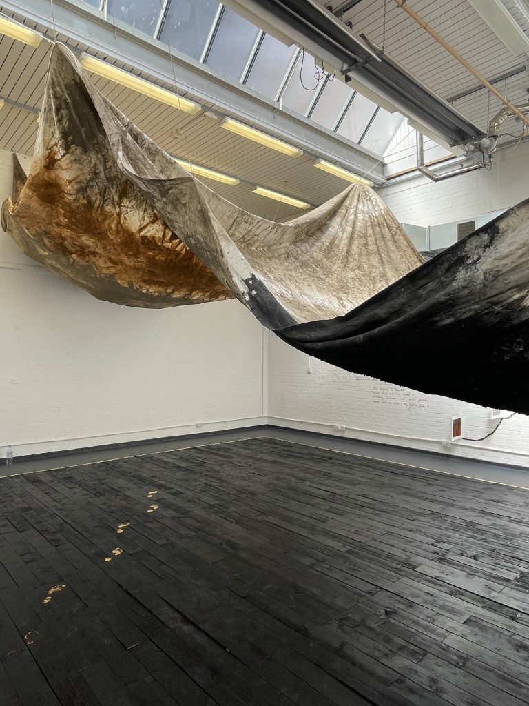 A large space with a floor make of charred wooden boards. Suspended from a slanted part of the ceiling is a large canvas or tarpaulin-like piece of fabric, painted with what looks like mud and charcoal.