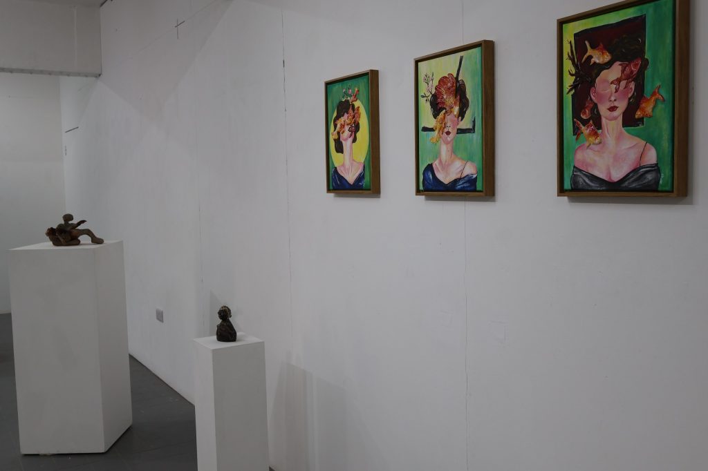 A white wall gallery with three portrait paintings hung in a row showing a woman's face and shoulders obscured by things including fish and plants