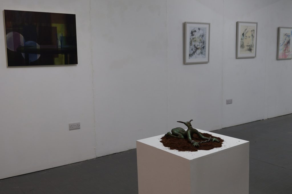 A small dark sculpture of a humanoid form with animal horns or ears, prostrated with wrists bound by rope, on a white plinth in the gallery