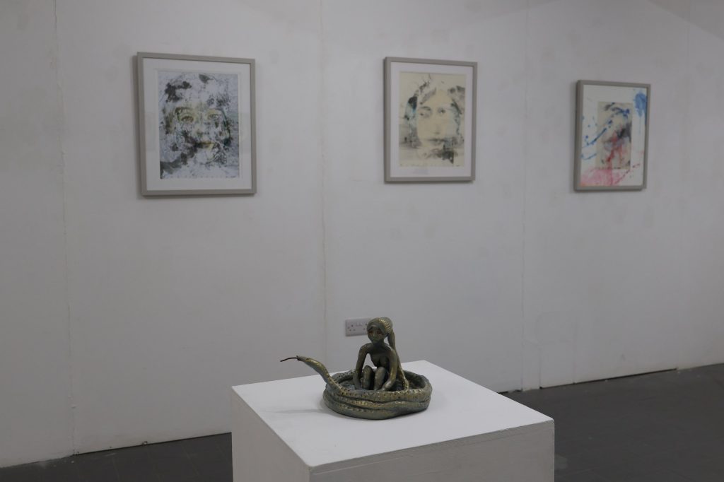 A small sculpture of a woman's sitting inside a coiled snake on a white plinth in the foreground with blurred framed images of human faces behind
