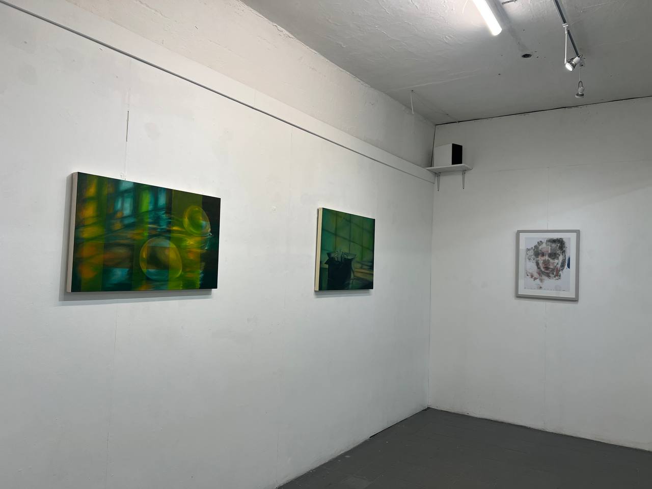 Three artworks hung in a row, two on the left are dark green paintings with spots of luminous light in grid shapes, and on the right an obscured greyscale human face