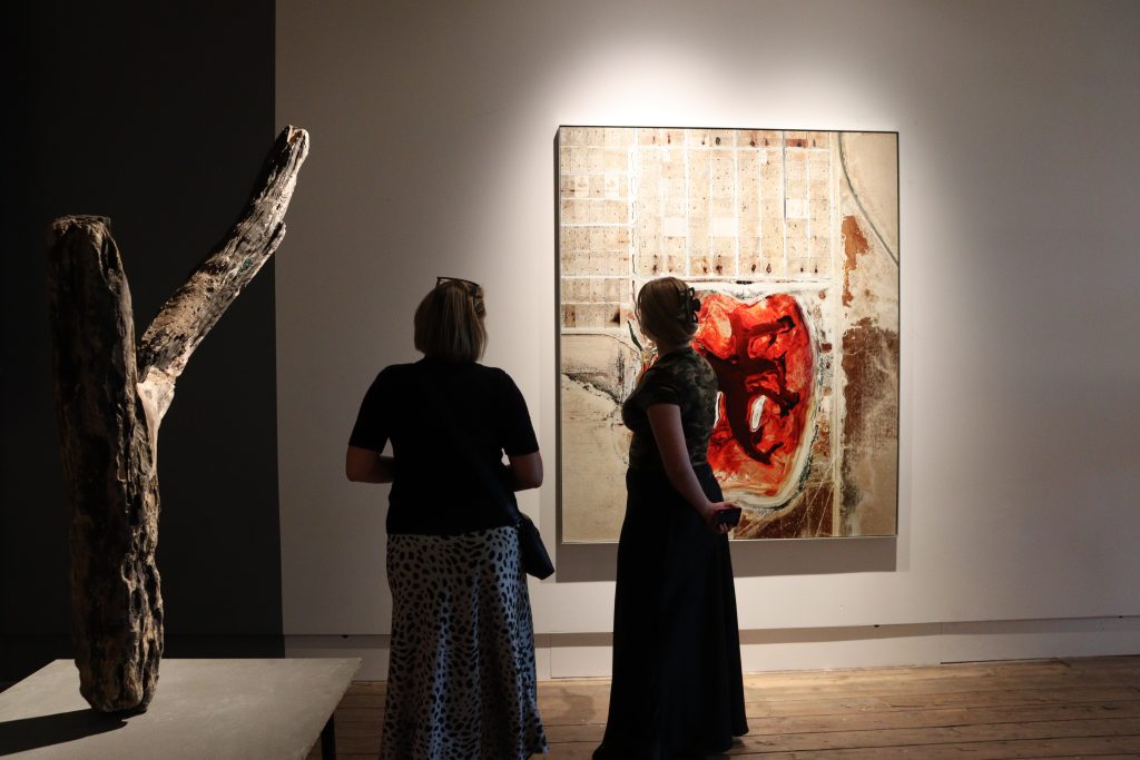 Two viewers look at a large, framed work hanging on a white wall, it is browns and beige with a huge dark red human heart shaped section in the middle