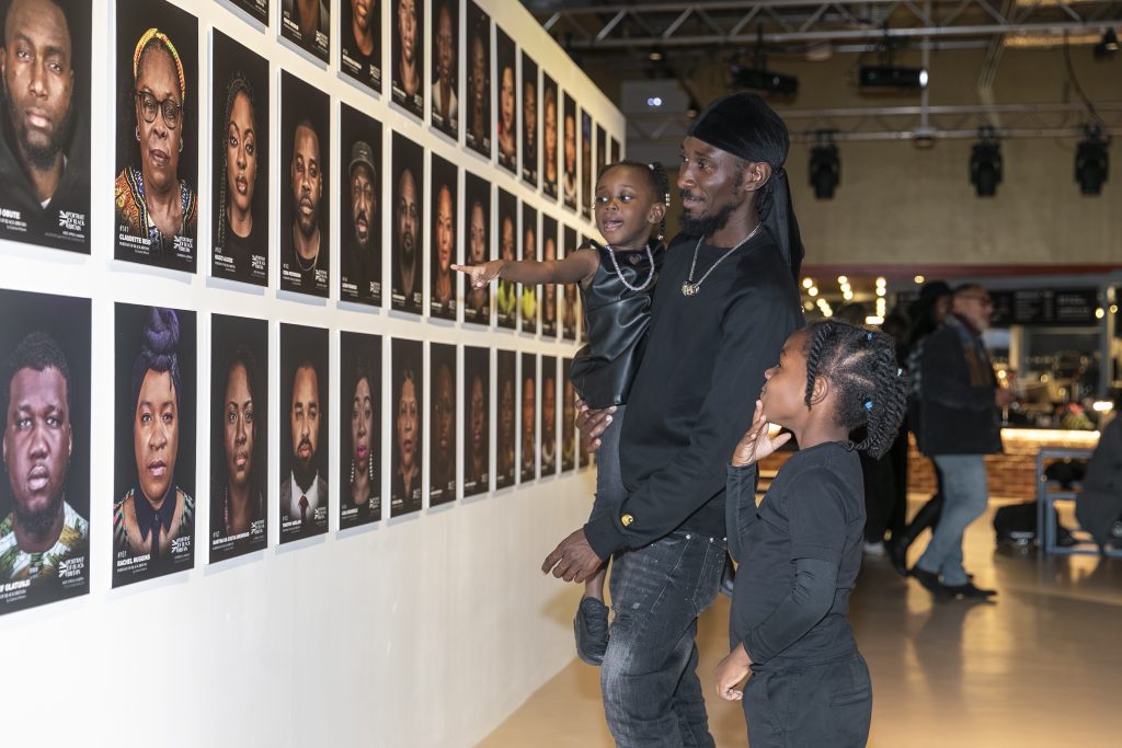 A Black man, with his two children, observe photos of Black people displayed in rows on a gallery wall