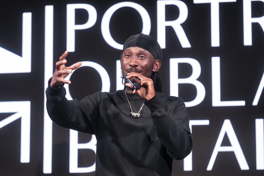 A Black Man speaks into a microphone. He is dressed in black and wears a black durag.
