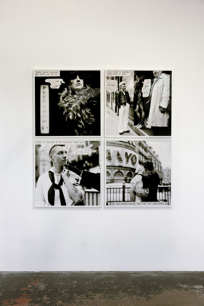 Four monochrome photographs in white frames are hung in a grid on a white gallery wall. They feature a sailor and a character in drag.