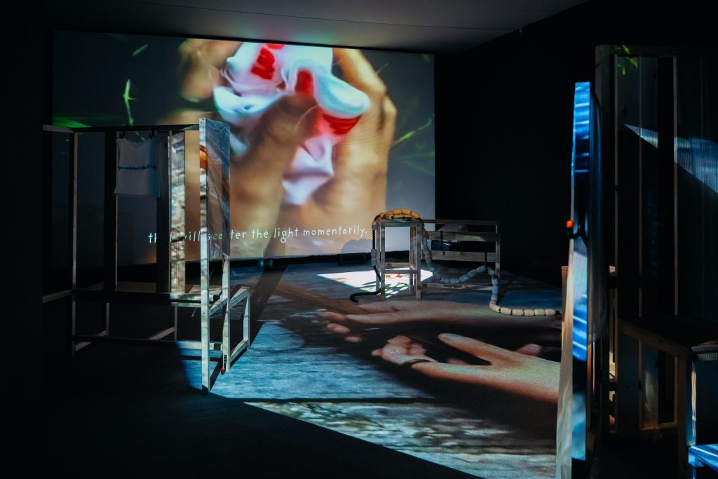 A darkened space in which a video is projected against one wall. It shows a still of two hands clutching something white and red - perhaps a piece of fabric. Wooden structures are dotted around the space in front, at times obstructing and distorting the projected image.
