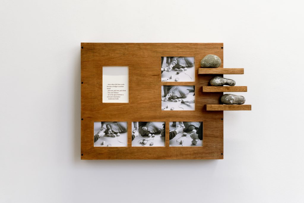 A wooden frame in which five small monochrome photos are inlaid, as well as a piece of text that is too small to read. Three small shelves on the right had side, which each have a stone sitting on them.