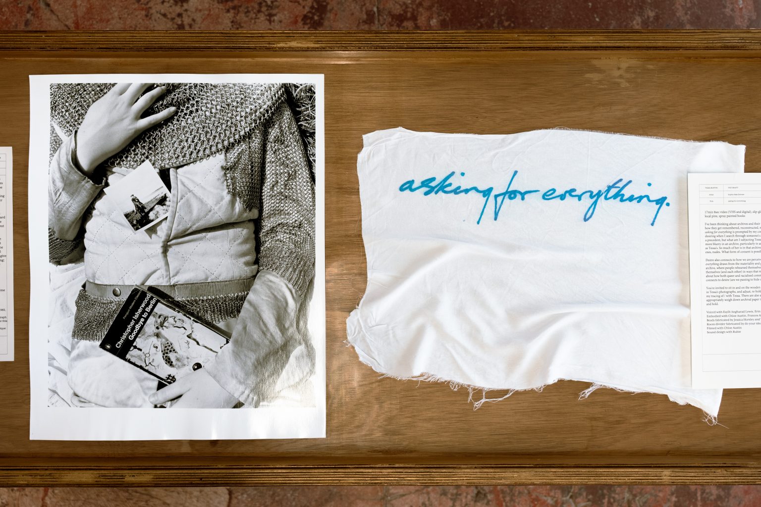 A monochrome photograph sits inside a wooden vitrine, next to it is a piece of white cloth with text in blue which reads 'asking for everything'.