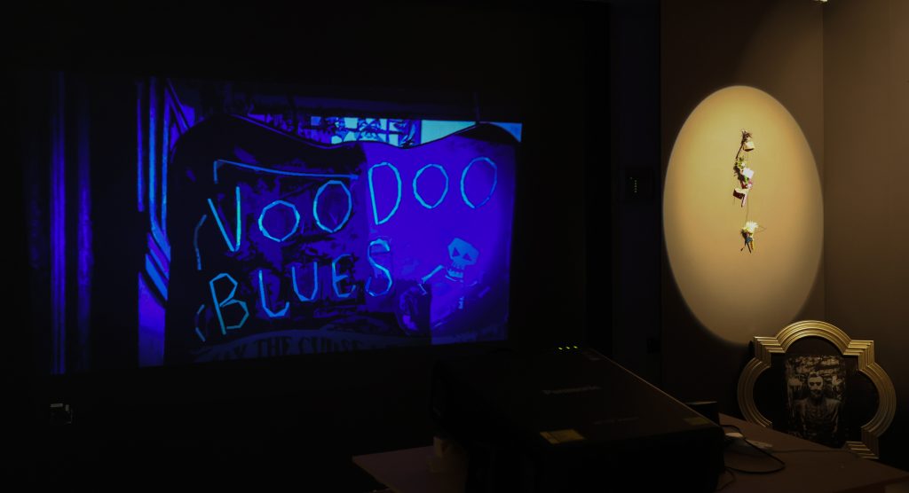 A darkened room in which a projected image in blue hues says 'Voodoo Blues' and other small objects are shown alongside.