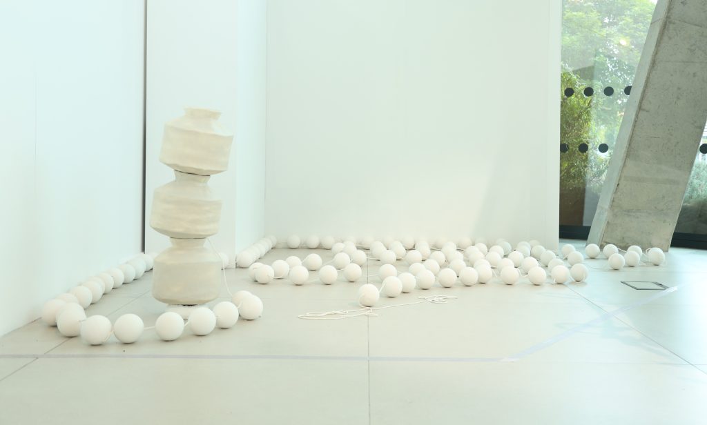 A white clay sculpture of an oversized string of beads curls around itself on the gallery floor. Three white clay vessels, resembling water carriers, are stacked on top of each other.
